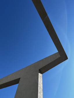 A large, abstract concrete structure with sharp angles and straight edges extends into a clear blue sky. The geometric shape creates a minimalist and modern aesthetic.