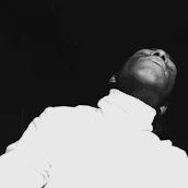 A person is leaning back with their head tilted upwards, wearing a white turtleneck against a black background. The image is high-contrast, emphasizing the contours and shadows on their face and neck.