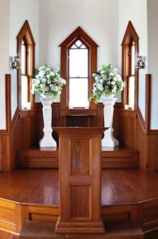 A wooden podium is centered in front of a small, elevated platform with large floral arrangements on white pedestals on either side. The area is framed by tall, narrow windows with wooden trim, creating a symmetrical and elegant setting.