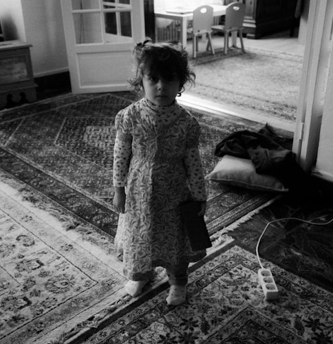 A young child stands in a room with ornate patterned rugs and a partially open door leading to another area with children's furniture. The child is wearing a patterned dress and holding an object. The setting feels domestic, with a power strip on the floor and a cushion nearby.