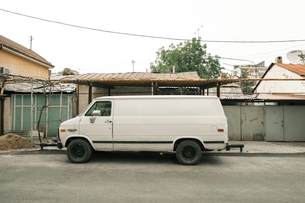 A white van is parked on a street in front of a series of garages or storage units. The scene includes utility poles with wires and a background with trees and greenery. The overall setting gives an urban or suburban feel, with flat, muted colors and a slightly overcast sky.