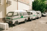 Three white vans with green and blue stripes are parked closely together on a gravel surface next to a building with air conditioning units. The vans have text on the side, which may indicate they are part of an official service or fleet. In the background, there are leafy green trees, and another vehicle is parked nearby.
