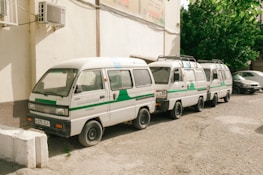Three white vans with green and blue stripes are parked closely together on a gravel surface next to a building with air conditioning units. The vans have text on the side, which may indicate they are part of an official service or fleet. In the background, there are leafy green trees, and another vehicle is parked nearby.