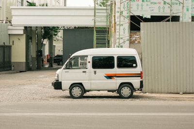 A white van parked beside a construction site with exposed scaffolding and building materials. The van has a stripe design in blue, orange, and red running along its side. The area around the van is unpaved, with gravel spread on the ground. Adjacent to the parked van, a large construction barrier is visible, and in the background, the framework of the building can be seen partially completed with insulation material covering parts.