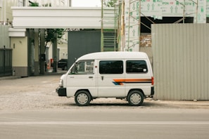 A white van parked beside a construction site with exposed scaffolding and building materials. The van has a stripe design in blue, orange, and red running along its side. The area around the van is unpaved, with gravel spread on the ground. Adjacent to the parked van, a large construction barrier is visible, and in the background, the framework of the building can be seen partially completed with insulation material covering parts.