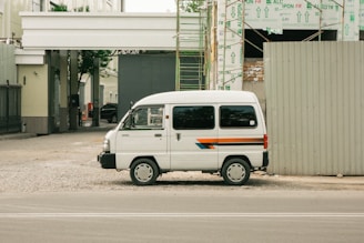 A white van parked beside a construction site with exposed scaffolding and building materials. The van has a stripe design in blue, orange, and red running along its side. The area around the van is unpaved, with gravel spread on the ground. Adjacent to the parked van, a large construction barrier is visible, and in the background, the framework of the building can be seen partially completed with insulation material covering parts.