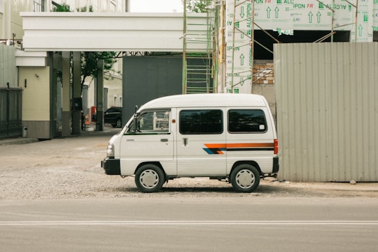A white van parked beside a construction site with exposed scaffolding and building materials. The van has a stripe design in blue, orange, and red running along its side. The area around the van is unpaved, with gravel spread on the ground. Adjacent to the parked van, a large construction barrier is visible, and in the background, the framework of the building can be seen partially completed with insulation material covering parts.
