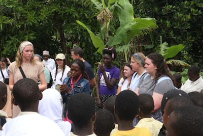 A diverse group of people stand outdoors surrounded by lush green vegetation. Some individuals appear to be engaged in discussion or listening attentively. The setting suggests a gathering or educational event, possibly involving cultural exchange.