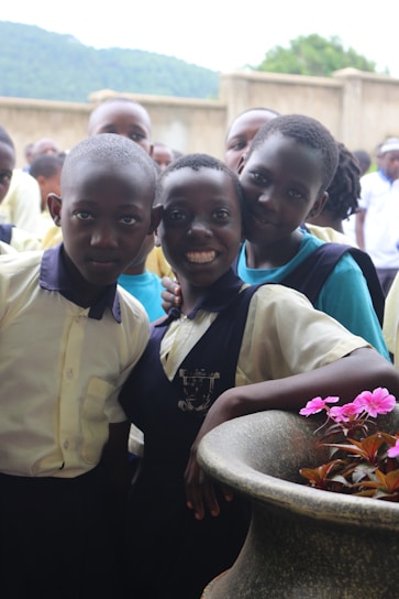 A group of smiling children is gathered in a courtyard area, with one child leaning on a large stone planter with bright pink flowers. They are wearing school uniforms and appear cheerful.