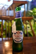 A green glass bottle of Peroni Nastro Azzurro beer sits on a wooden table. The label is prominently displayed with blue and red text. The background features an out-of-focus indoor setting with wooden furniture and green plants.