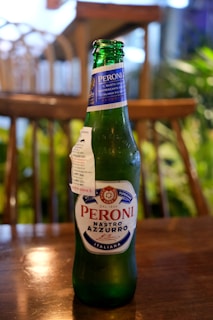 A green glass bottle of Peroni Nastro Azzurro beer sits on a wooden table. The label is prominently displayed with blue and red text. The background features an out-of-focus indoor setting with wooden furniture and green plants.