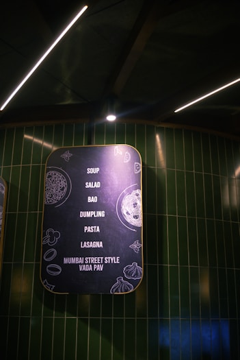 A stylishly designed menu board with a matte black background and yellow border features a list of food items including soup, salad, bao, dumpling, pasta, lasagna, and Mumbai street style vada pav. The board is decorated with minimalistic line drawings of food ingredients and illuminated by overhead lighting against a backdrop of dark green tiled walls.