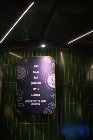 A stylishly designed menu board with a matte black background and yellow border features a list of food items including soup, salad, bao, dumpling, pasta, lasagna, and Mumbai street style vada pav. The board is decorated with minimalistic line drawings of food ingredients and illuminated by overhead lighting against a backdrop of dark green tiled walls.