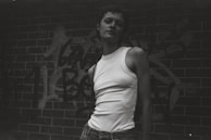 A monochrome shot of a model in slate grey joggers and a white tee, leaning against a graffiti-covered wall.