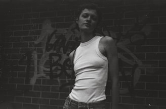 A monochrome shot of a model in slate grey joggers and a white tee, leaning against a graffiti-covered wall.