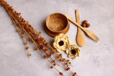 A minimalist arrangement featuring a wooden bowl, spoon, and fork on a textured surface. Nearby, cream-colored artificial flowers with dark centers add a decorative touch. Brown woven balls and dried brown stems extend across the image, creating an earthy composition.