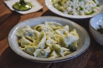 A plate of homemade dumplings with a green filling inside sits on a rustic wooden table, accompanied by a bowl of mixed rice and peas in the background. The scene is set with natural lighting, highlighting the texture of the wooden surface and the food.