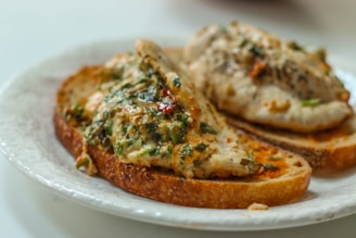 Two slices of toasted bread topped with grilled chicken breasts garnished with herbs and spices, served on a white plate. The herbs appear fresh with hints of green and red pepper flakes visible.