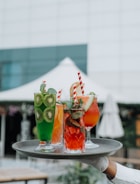 A tray holds an assortment of colorful drinks in glasses garnished with fruits and mint leaves. Each beverage is uniquely vibrant, featuring layered colors and striped straws. The background shows a blurred outdoor setting with a tent and greenery.