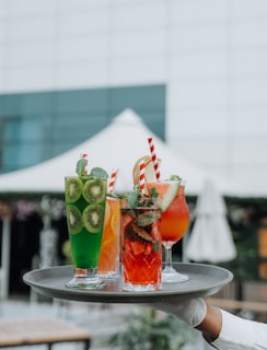 A tray holds an assortment of colorful drinks in glasses garnished with fruits and mint leaves. Each beverage is uniquely vibrant, featuring layered colors and striped straws. The background shows a blurred outdoor setting with a tent and greenery.