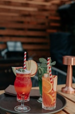 A selection of colorful cocktails is artfully arranged on a round tray. Each drink is garnished with fresh fruits like apple slices, kiwi, watermelon, orange slices, and topped with mint leaves. The drinks have vibrant colors such as red, green, and orange, and are served in various types of glassware. The setting has a warm, wooden background that gives a cozy ambiance.