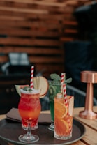 A selection of colorful cocktails is artfully arranged on a round tray. Each drink is garnished with fresh fruits like apple slices, kiwi, watermelon, orange slices, and topped with mint leaves. The drinks have vibrant colors such as red, green, and orange, and are served in various types of glassware. The setting has a warm, wooden background that gives a cozy ambiance.
