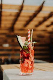 A tall glass filled with a bright pink drink, garnished with a slice of watermelon and mint leaves. The drink also contains ice cubes and is accompanied by two straws, one striped and one plain. The background consists of a wooden structure, creating a warm ambiance.