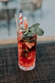 A tall glass filled with a vibrant red beverage, garnished with mint leaves and a slice of watermelon. The glass features an intricate diamond pattern and contains two straws, one paper with red and white stripes and another bamboo straw. The drink is placed on a woven mat surface with a blurred background.