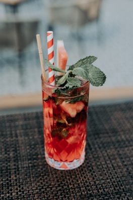 A tall glass filled with a vibrant red beverage, garnished with mint leaves and a slice of watermelon. The glass features an intricate diamond pattern and contains two straws, one paper with red and white stripes and another bamboo straw. The drink is placed on a woven mat surface with a blurred background.