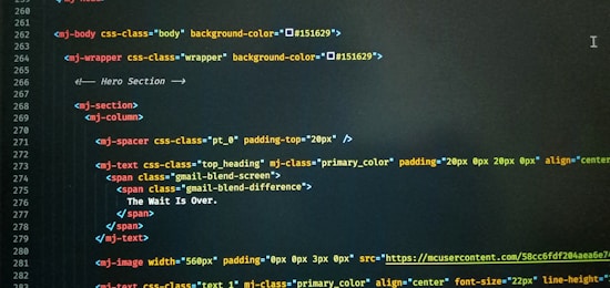 A computer screen displaying code with HTML-like tags and inline CSS styles. The code featured includes elements such as mj-body, mj-wrapper, and mj-text with specific styling for background colors, padding, alignment, and text content. Notable text within the code includes 'The Wait Is Over.'