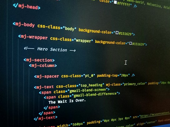 HTML code displayed on a computer screen, featuring elements such as 'mj-body', 'mj-wrapper', and 'mj-text'. The code includes CSS classes for styling purposes, indicating colors with hex codes like '#151629'. A comment suggests this section is the 'Hero Section'.