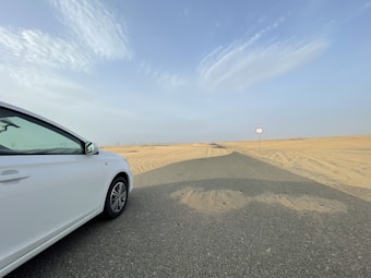 A white car is partially visible on the left side of a road that stretches into a desert. The road is bordered by sand dunes under a clear, bright sky. A circular speed limit sign is visible on the right side. The scene is expansive with an open, empty road and vast desert landscape in the background.
