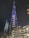 A towering skyscraper illuminated at night with vibrant blue and white lights, featuring the word 'EMAAR' prominently displayed. The building is surrounded by other illuminated modern structures and a palm tree.