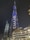 A towering skyscraper illuminated at night with vibrant blue and white lights, featuring the word 'EMAAR' prominently displayed. The building is surrounded by other illuminated modern structures and a palm tree.