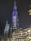 A towering skyscraper illuminated at night with vibrant blue and white lights, featuring the word 'EMAAR' prominently displayed. The building is surrounded by other illuminated modern structures and a palm tree.