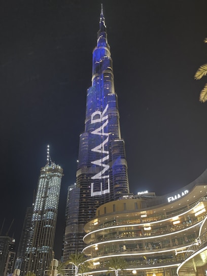 A towering skyscraper illuminated at night with vibrant blue and white lights, featuring the word 'EMAAR' prominently displayed. The building is surrounded by other illuminated modern structures and a palm tree.