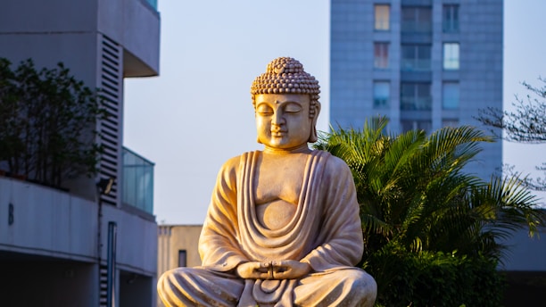 A stone statue of Buddha sits peacefully in a meditative pose amidst a modern urban environment. The statue is illuminated by warm sunlight, casting gentle shadows. Behind the Buddha, there is lush green foliage and a tall building, giving a contrast between nature and urban life.