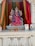 A colorful statue of a deity with multiple arms, dressed in an ornate pink dress and detailed crown, is placed on an intricately carved pedestal. The background features vibrant red, orange, and green drapery. Flowers are scattered on the platform in front, accompanied by a metallic donation box.