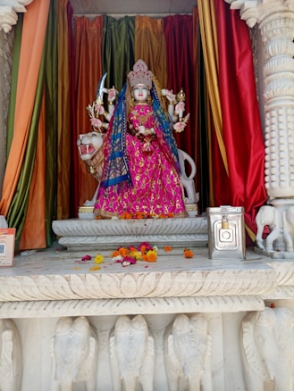A colorful statue of a deity with multiple arms, dressed in an ornate pink dress and detailed crown, is placed on an intricately carved pedestal. The background features vibrant red, orange, and green drapery. Flowers are scattered on the platform in front, accompanied by a metallic donation box.