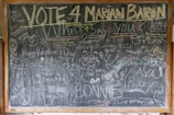 A wooden-framed blackboard is filled with chalk writings and drawings. Various messages such as 'Vote 4 Marian Baron', 'Happy Valentine's Day', 'Peace and Love', and 'I love you!' are inscribed amidst doodles, smiley faces, and peace symbols. The board also includes random names, hearts, and phrases in different sizes and orientations.