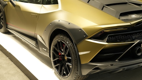 A luxury sports car with a sleek design, featuring a matte gold finish and black accents. The car has a distinctive rear section with aerodynamic elements and large black alloy wheels. The tire treads are distinctly visible, and a V10 badge is spotted near the engine cover.