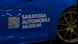 A blue car with a QR code and the text 'Saratoga Automobile Museum' displayed on its side. The wheel is visible, featuring golden rims with the label 'GT4' and 'Porsche'.