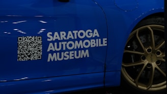 A blue car with a QR code and the text 'Saratoga Automobile Museum' displayed on its side. The wheel is visible, featuring golden rims with the label 'GT4' and 'Porsche'.