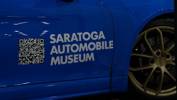 A blue car with a QR code and the text 'Saratoga Automobile Museum' displayed on its side. The wheel is visible, featuring golden rims with the label 'GT4' and 'Porsche'.