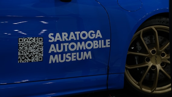 A blue car with a QR code and the text 'Saratoga Automobile Museum' displayed on its side. The wheel is visible, featuring golden rims with the label 'GT4' and 'Porsche'.