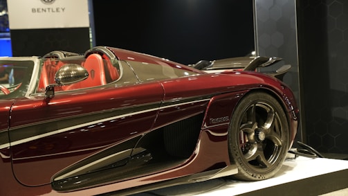 A luxury sports car with a sleek and aerodynamic design displayed in a showroom. The car features a glossy maroon color with black and chrome accents. The interior is visible, showcasing red seats and a high-performance steering wheel. Carbon fiber elements are visible on the body, emphasizing its sporty nature. The image includes a partially visible sign with the Bentley logo in the background.