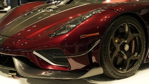 A close-up view of the front section of a luxury sports car. The car features a sleek, aerodynamic design with a deep burgundy and black carbon fiber exterior. The front wheel is visible, showcasing high-performance tires and intricate rim details. Reflections of overhead lights can be seen on the car's surface, enhancing its glossy finish.