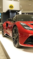 A sleek, shiny red sports car showcased indoors, positioned on a white platform. The car features aerodynamic curves, prominent black rims, and a low profile, indicative of a high-performance vehicle. In the background, a yellow and black logo is displayed on a stand.