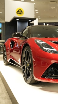 A sleek, shiny red sports car showcased indoors, positioned on a white platform. The car features aerodynamic curves, prominent black rims, and a low profile, indicative of a high-performance vehicle. In the background, a yellow and black logo is displayed on a stand.