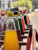 A row of colorful glass bottles filled with various liquids is arranged in a wooden crate. The bottles have different colored caps, including yellow, red, and green, and contain liquids in shades of yellow, red, green, and orange. The setting appears outdoors, with a blurred background of foliage and a street.